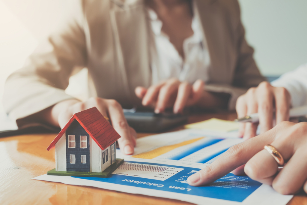 A small house model on a table with real estate documents and people discussing.