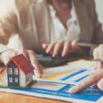A small house model on a table with real estate documents and people discussing.