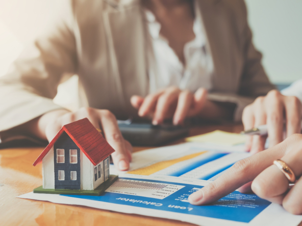 A small house model on a table with real estate documents and people discussing.